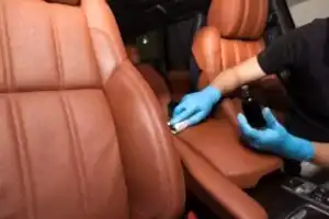 Applying a nano-ceramic coating on leather of the car seat brown upholstery by a worker in blue gloves with a sponge and bottle